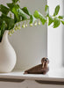 A Spring Copenhagen Wally sculpture with large, round eyes and a smiling mouth sits on a white surface, next to a vase holding white lily flowers. Bright natural light illuminates the scene.