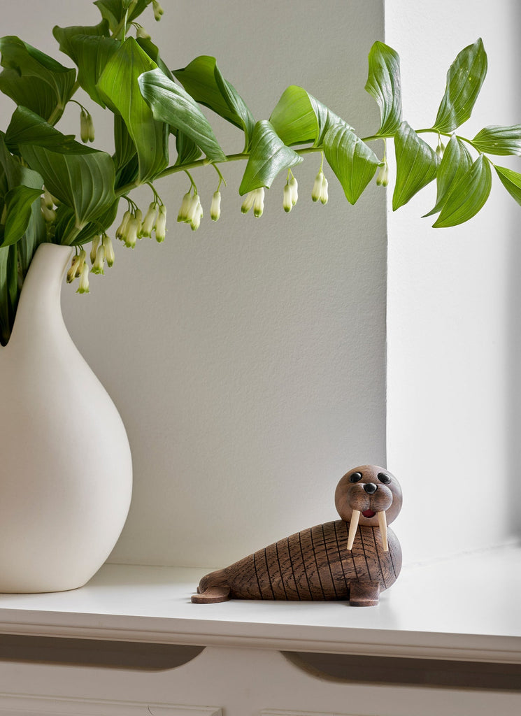 A Spring Copenhagen Wally sculpture with large, round eyes and a smiling mouth sits on a white surface, next to a vase holding white lily flowers. Bright natural light illuminates the scene.