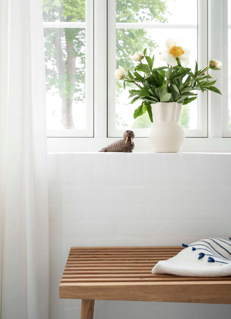 A bright room with a large window showing green trees outside. A ceramic vase with yellow and white flowers is on the windowsill, next to a small handmade wooden Spring Copenhagen Wally in the form of.