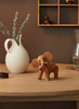 A Spring Copenhagen Ollie - Elephant, crafted from FSC Oak, stands on a table in a warmly lit room with a white vase, dried plants, and other decorative items on shelves.