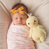 Newborn baby girl wearing a yellow bow headband, swaddled in a floral pink blanket, sleeping beside a Cuddle + Kind Baby Duckling toy made from Peruvian cotton yarn.