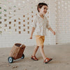 A young boy smiling as he walks and pulls a small hand-woven Olli Ella Rattan Luggy with wheels, dressed in a patterned shirt and orange shorts, against a backdrop of a white brick.