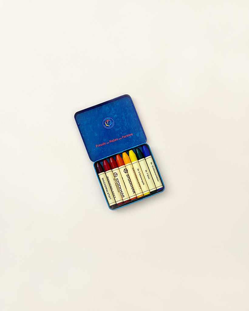 A set of Stockmar Wax Stick Crayons Waldorf Tin Case - 8 Assorted arranged neatly in an open blue metal case, positioned on a plain light background. Each crayon is labeled with a different color name.