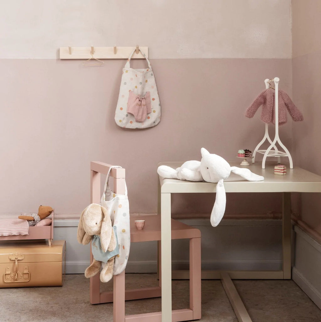 A cozy children's room corner featuring a wooden desk with toys like a Maileg Bunny Holly and a plush elephant, shelves with books, and a pastel pink wall. A hanging polka dot bag adorned with