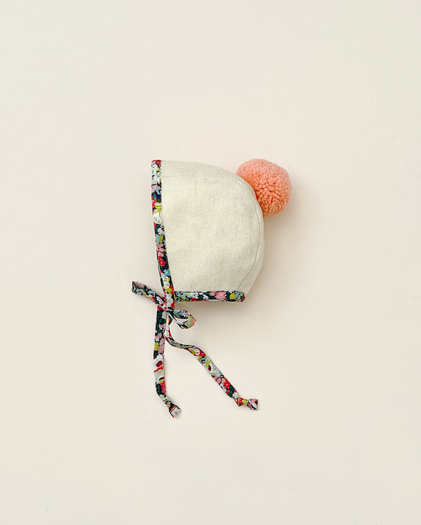A Briar Baby Wild Poppy Pom Bonnet with a Liberty of London cotton floral print tie and a soft orange pompom attached, set against a plain, light beige background.