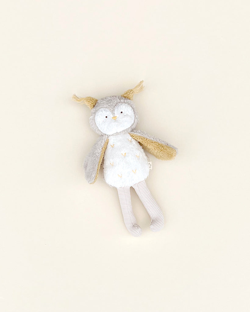 A soft plush toy with a bunny design, featuring a grey and white color scheme, lying flat on a pale yellow background. The Owl Stuffed Animal has long ears, embroidered details, and a striped pattern on its belly.