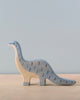 A Holztiger Brontosaurus stands on a light wooden surface. The handcrafted wood dinosaur is painted in light blue and beige colors, with blue crescent-shaped spots along its body. Made in Europe by HOLZTIGER figures, the background is a plain, light blue wall.