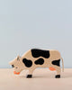 A Holztiger Grazing Black Cow with black spots and a tan body stands on a light tan surface against a pale blue background. The handcrafted wood toy, made in Europe, is slightly bent forward as if grazing.