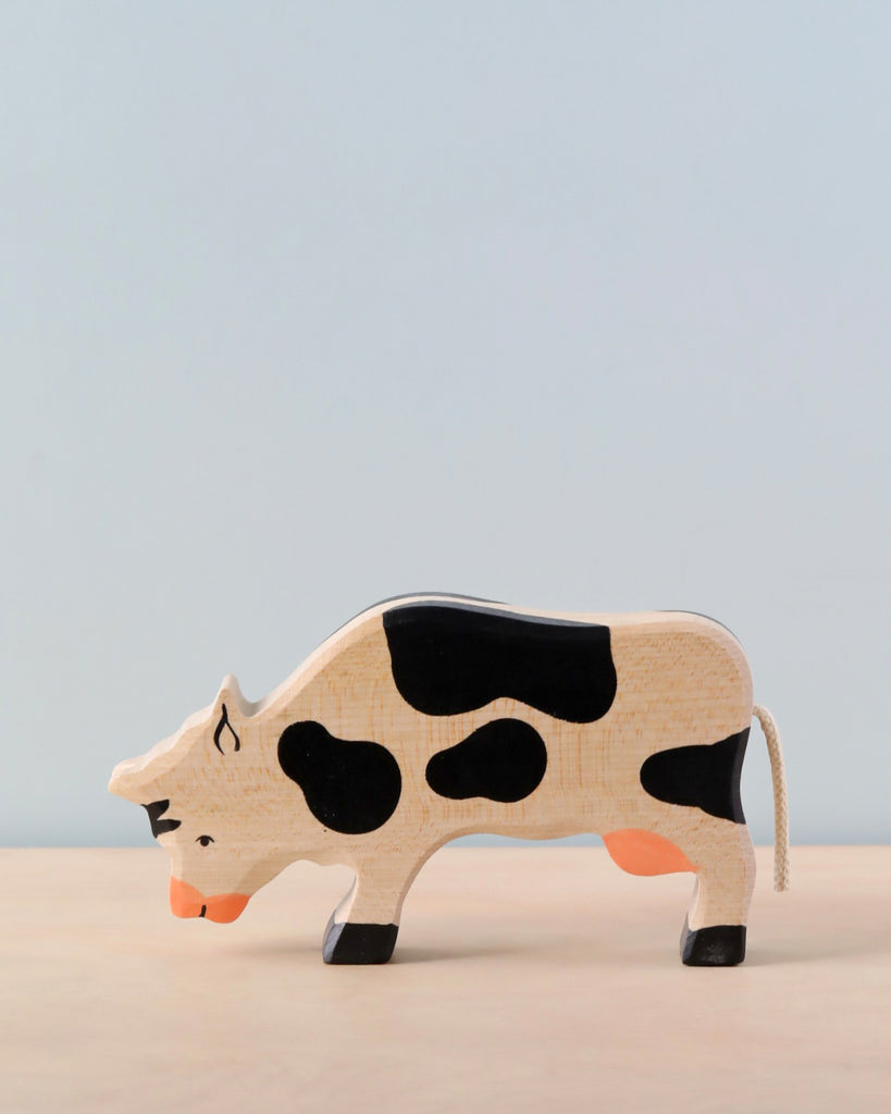 A Holztiger Grazing Black Cow with black spots and a tan body stands on a light tan surface against a pale blue background. The handcrafted wood toy, made in Europe, is slightly bent forward as if grazing.