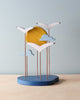 A minimalist wooden kinetic sculpture, hand-made in Greece, featuring four white seabirds with outstretched wings, set against a round yellow backdrop evoking a sunset, mounted on a circular blue base
