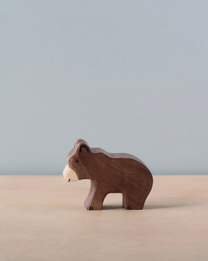 The Holztiger Small Brown Bear, a handcrafted wooden figurine, stands on a light wooden surface with a plain, pale blue background. Carved with simple features and stained brown with a lighter muzzle, this European-made piece exudes rustic charm.