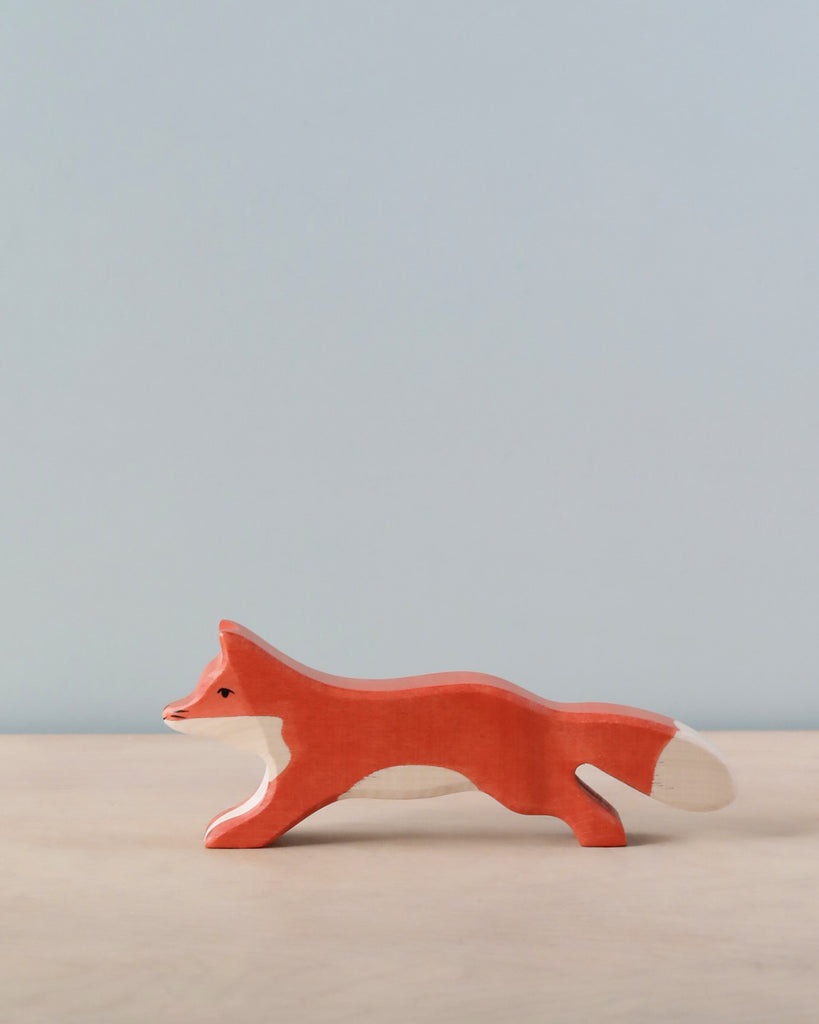 A handcrafted wooden toy fox stands on a light wood surface against a pale blue wall. The fox, part of the Holztiger Running Fox collection, has an orange body with white accents on its face, belly, and tail, and is captured in mid-stride. Made in Europe.