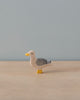 A small wooden seagull figurine with a white head, yellow beak and feet, gray body, and black and white tail feathers stands on a simple light brown wooden surface against a plain pale blue background. Like other Holztiger Seagull figures, it reflects the timeless charm of handcrafted wooden toys made in Europe.