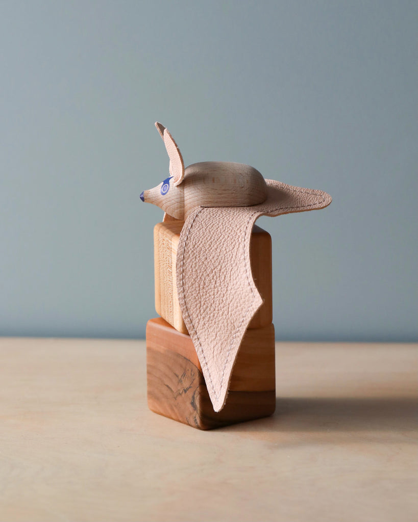 A handmade wooden bat, crafted with textural details and large flapping ears made from leather, stands on a square wooden base against a neutral background.