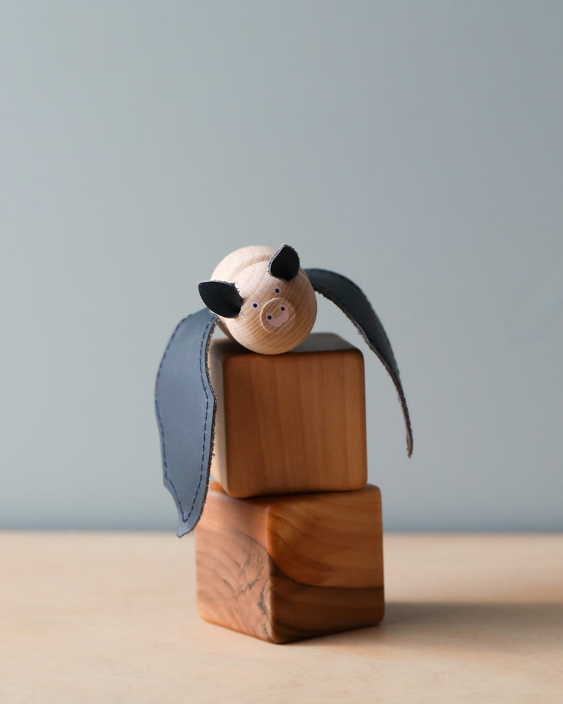 A handmade wooden bat with fabric ears and leather wings perched atop a small, stacked wooden block, set against a soft blue background.