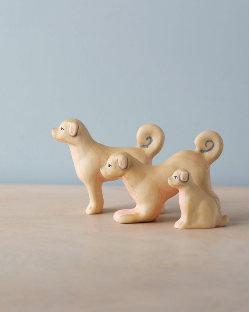 The Handmade Wooden Dog Family includes three small wooden dog figures of varying sizes, each with curled tails and simple facial features, displayed on a light wooden surface against a pale blue background.