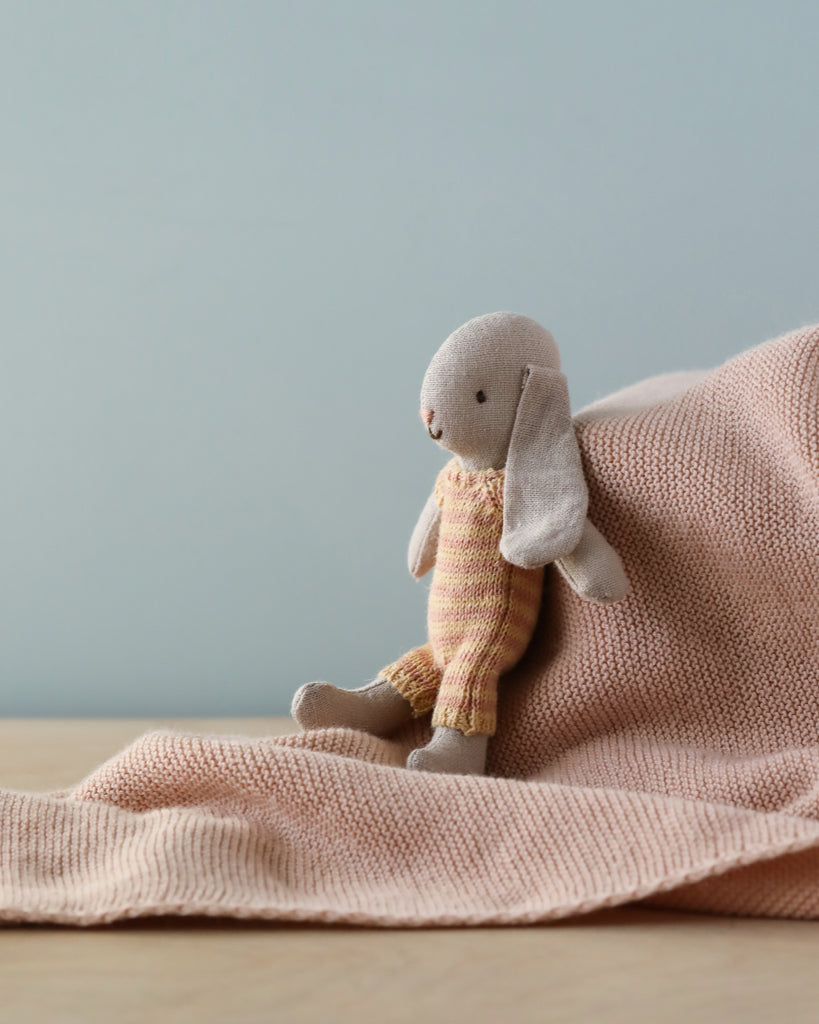 A Maileg Micro Bunny plush toy sitting on a pink knitted blanket against a soft blue background. The toy has a thoughtful expression, looking off to the side.