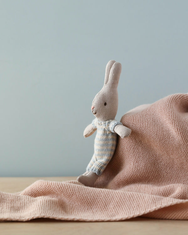 A Maileg Micro Rabbit dressed in a striped knitted suit, sitting against a wooden surface with a gray background.