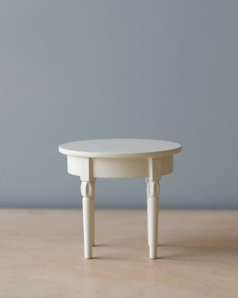 A small, round, cream-colored Maileg | Mini Side Table with a simple, elegant design, featuring tapered legs. The table is set against a soft gray background.