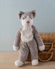 A Senger Naturwelt Stuffed Animal - Cat, handmade in Germany, with grey fur and a white face/belly stands upright against a basket on a wooden table, with a pale blue background.