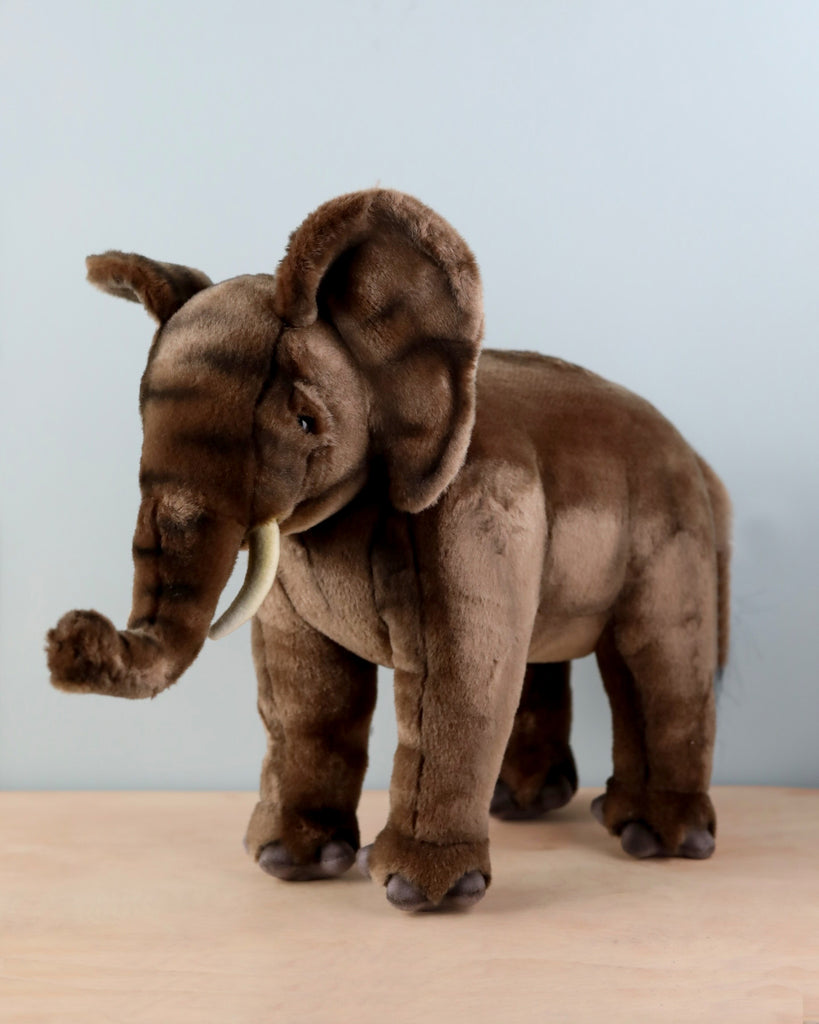 A unique Elephant Stuffed Animal with soft brown fur, detailed tusks, and large floppy ears stands on a wooden surface against a light grey background. The realistic plush animal is depicted with its trunk slightly curved upwards.