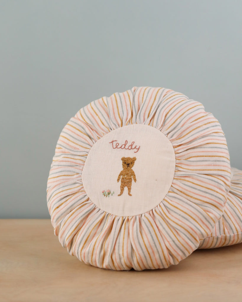 A round Maileg Cushion, Small with a beige and white striped border. The center features an embroidered design of a small teddy bear with the word "teddy" above it, set against a white background.