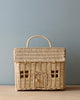 The Olli Ella Rattan Casa Clutch is a wicker basket shaped like a toy house, featuring a handle on top, woven pattern, faux windows, and door. It sits elegantly on a wooden surface against a plain light blue background.