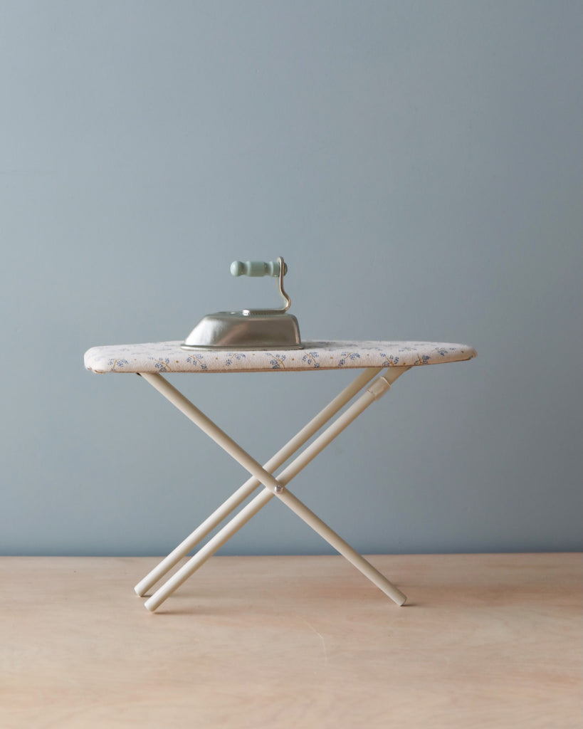 A Maileg | Miniature Iron & Ironing Board resting on a small ironing board with a floral cover, set against a plain gray wall and light wooden floor.