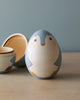 A handcrafted Hand Painted Hollow Wooden Easter Egg - Penguin, painted to resemble a cute, chubby bird, with one half of the egg opened to show the hollow inside, set on a wooden surface against a soft blue background.