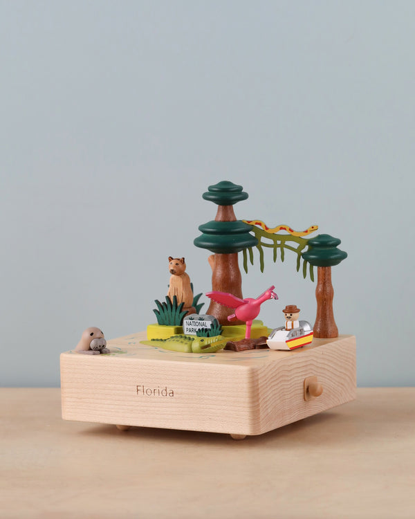 A Florida Wooden Music Box featuring miniature figures: a squirrel, a flamingo, a manatee, a boat, and palm trees, set against a neutral background.