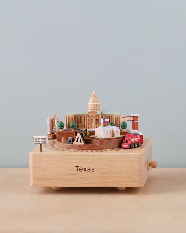 A Texas Wooden Music Box featuring cultural landmarks and icons of Texas, including the state capitol and a red truck, set against a plain background.