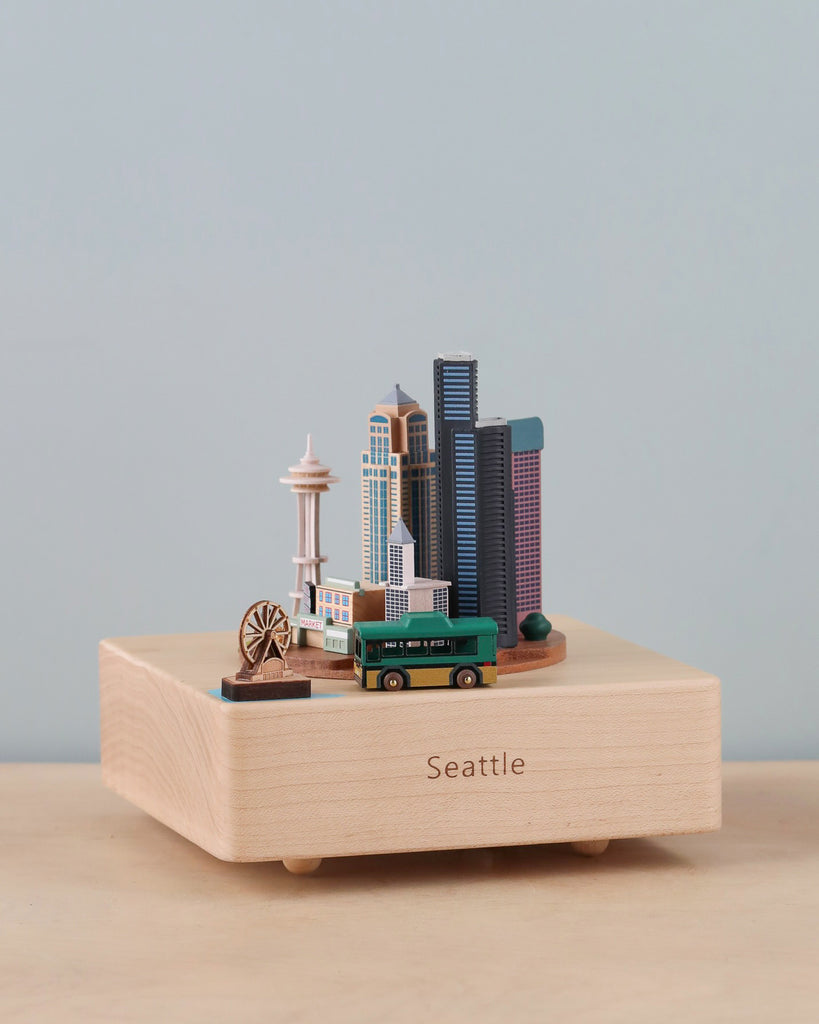 The Seattle Wooden Music Box is a small wooden model of Seattle showcasing miniature replicas of the Space Needle, Seattle Great Wheel, city buildings, and a bus, all mounted on a rectangular wooden base labeled "Seattle." This unique piece highlighting the city's culture and landmarks is the perfect gift or keepsake. The background is plain and light-colored.