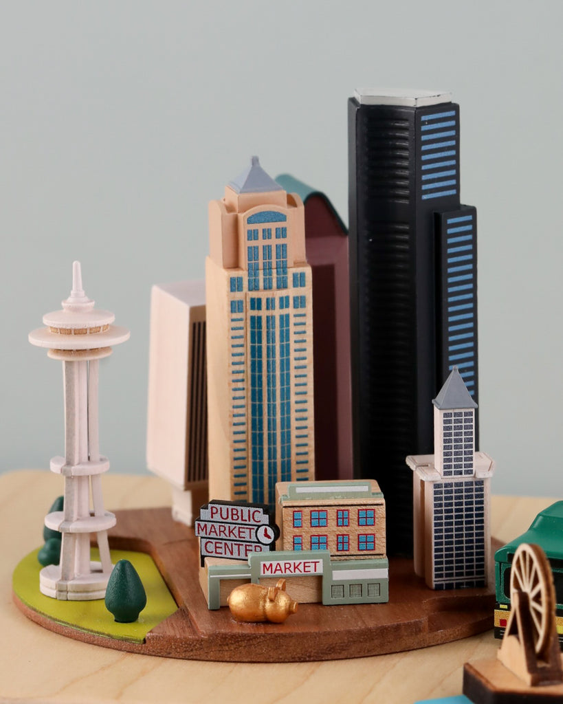 The Seattle Wooden Music Box showcases a delightful miniature model diorama featuring notable Seattle landmarks, including the iconic Space Needle, a "Public Market Center" sign, and several skyscrapers. The scene is intricately detailed with small trees and additional buildings, all elegantly set on a circular wooden base—capturing Seattle's unique culture and landmarks beautifully.