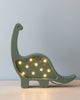 A decorative green Handmade Small Dinosaur Lamp with small round lights embedded along its side and back, resting on a solid pinewood surface against a plain light blue background.