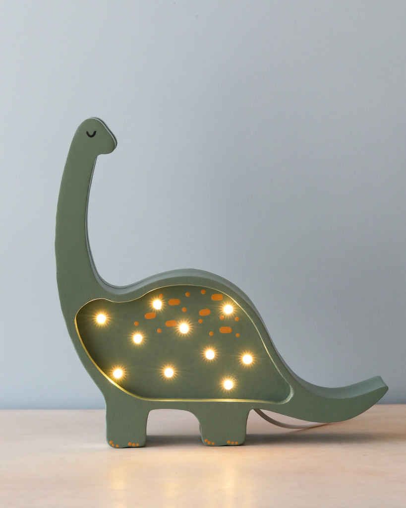 A decorative green Handmade Small Dinosaur Lamp with small round lights embedded along its side and back, resting on a solid pinewood surface against a plain light blue background.