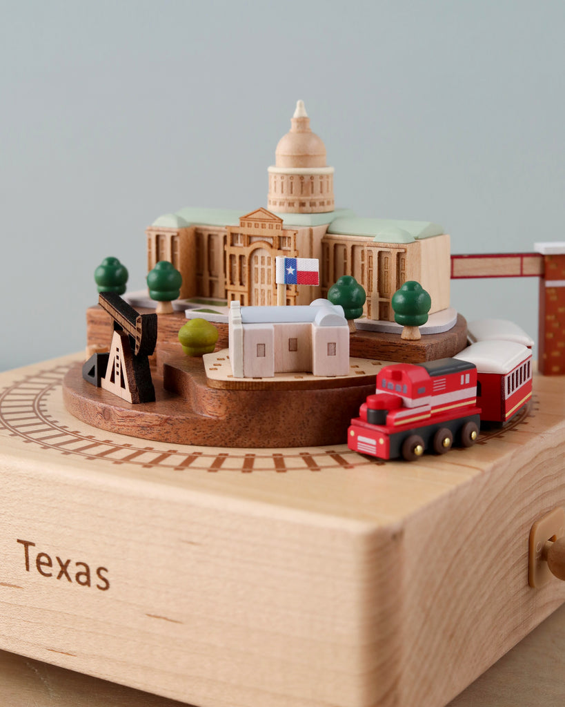 A detailed miniature model of the Texas Wooden Music Box cultural landmarks, including the state capitol, placed on a wooden base with a red and white train circling around. The base is labeled "Texas.