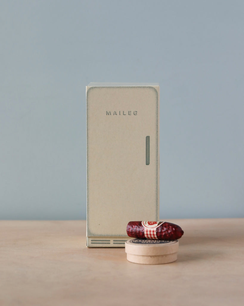 A small cardboard box styled like a vintage wardrobe stands on a muted blue background, with a red fabric mouse bed in front. The box is labeled "Maileg Cooler.