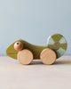 Wooden green chameleon with light blue stripes on tail, natural color wheels and eyes. Beige counter and light blue background. 