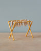 A small, yellow, tabletop Maileg Drying Rack, Mouse Size stands on a wooden surface. The rack has small wooden pegs clipped along its horizontal bars, giving it the appearance of a miniature laundry drying setup. The background is a plain, muted light blue wall.