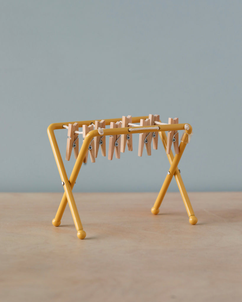 A small, yellow, tabletop Maileg Drying Rack, Mouse Size stands on a wooden surface. The rack has small wooden pegs clipped along its horizontal bars, giving it the appearance of a miniature laundry drying setup. The background is a plain, muted light blue wall.