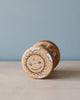 A handmade wooden rattle, crafted from sustainably harvested birch wood, with a smiling cartoon sun face drawn on it, resting on a light wood surface against a soft blue background.