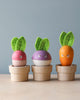 Three wooden toys resembling vegetables with smiling faces stand on a table in small wooden pots. From left to right, the toys are a red radish, a purple turnip, and an orange carrot, each with green felt leaves on top. Perfect for budding gardeners, this Stacking Wooden Veggies set features a light blue wall background.