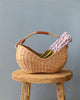 A Basket of Lavender Felt Flowers with a curved handle, containing a lilac knitted scarf and green beans, resting on a rustic wooden stool against a gray background.