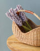 A hand-woven Basket of Lavender Felt Flowers with a curved handle holds a bouquet of purple lavender flowers. The basket is placed on a wooden surface against a pale blue background.