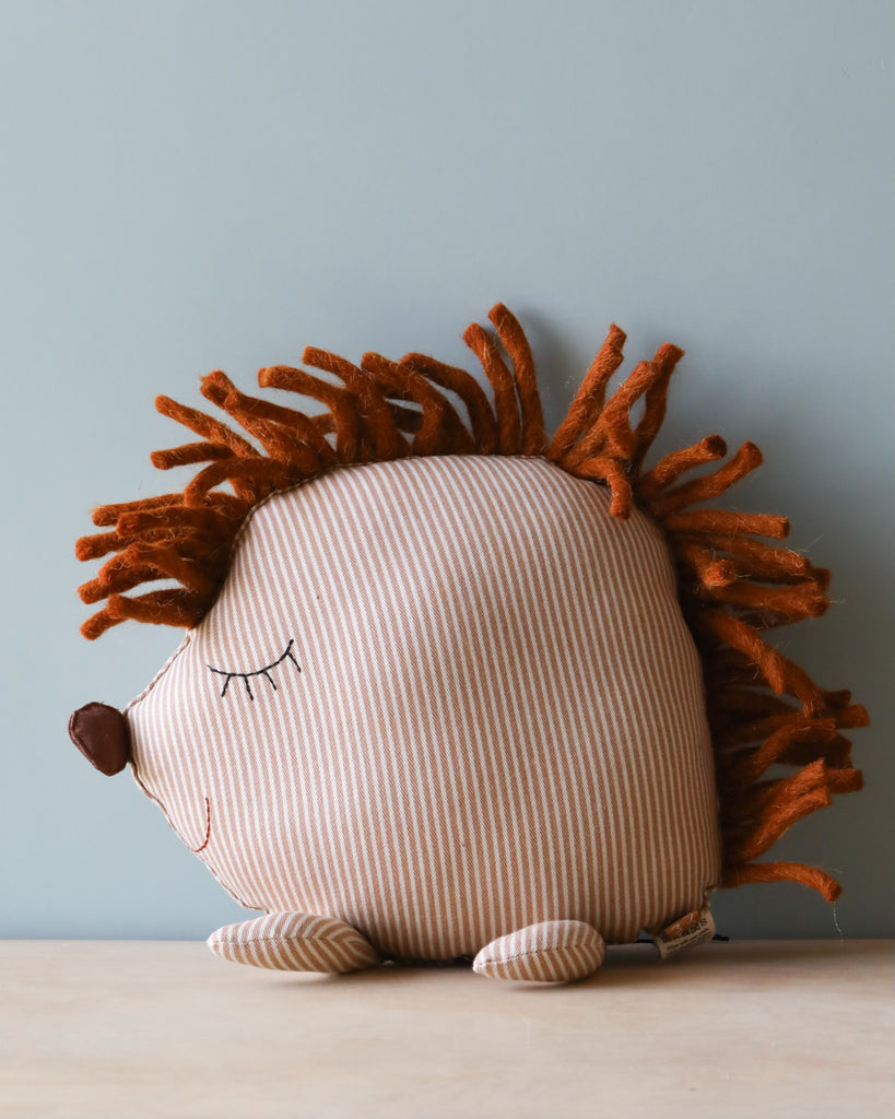 A cute, plush Hedgehog Cushion toy with a striped body and brown, textured fabric spikes sits against a light blue background. Its eyes are closed, and it has a small, brown nose.