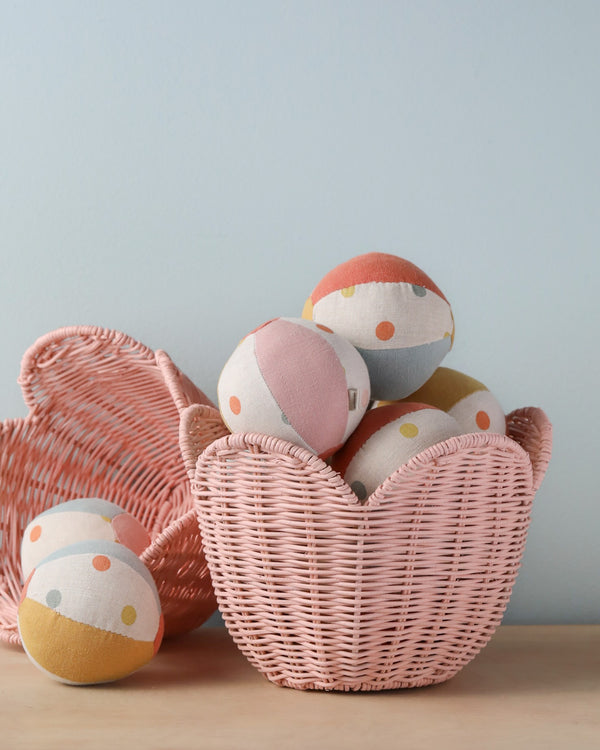 A pink woven basket shaped like a heart, filled with colorful polka-dotted fabric balls, next to another empty Olli Ella Rattan Lily Basket Set, on a wooden surface against a pale blue background.
