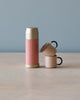 A pink Maileg | Miniature thermos and two matching cups with magnetic hands on a wooden table against a pale blue background.