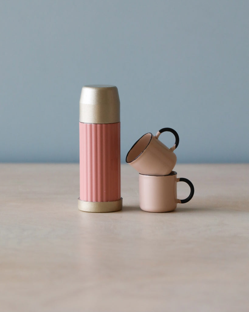 A pink Maileg | Miniature thermos and two matching cups with magnetic hands on a wooden table against a pale blue background.