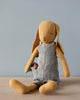 A Maileg Bunny Size 3, Dusty Yellow with long floppy ears sitting against a plain background, wearing a plaid dress made from soft natural linen and holding a tiny basket.
