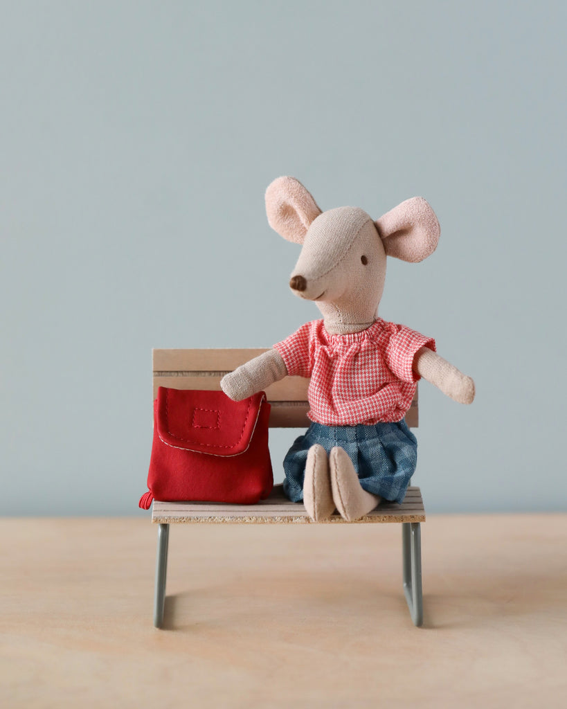 A Maileg Big Sister With Backpack - Red dressed in a red checkered shirt and denim shorts, sitting on a small bench with a red bag beside it, against a soft blue background.